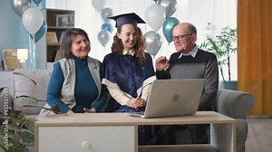 parents and female graduate happily clapping hands during graduation ceremony via video link on laptop while sitting at home on couch with balloons in background