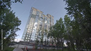 High-rise office tower in surrounding of greenery of urban park. Motion along narrow causeway towards building reflecting sky in windows